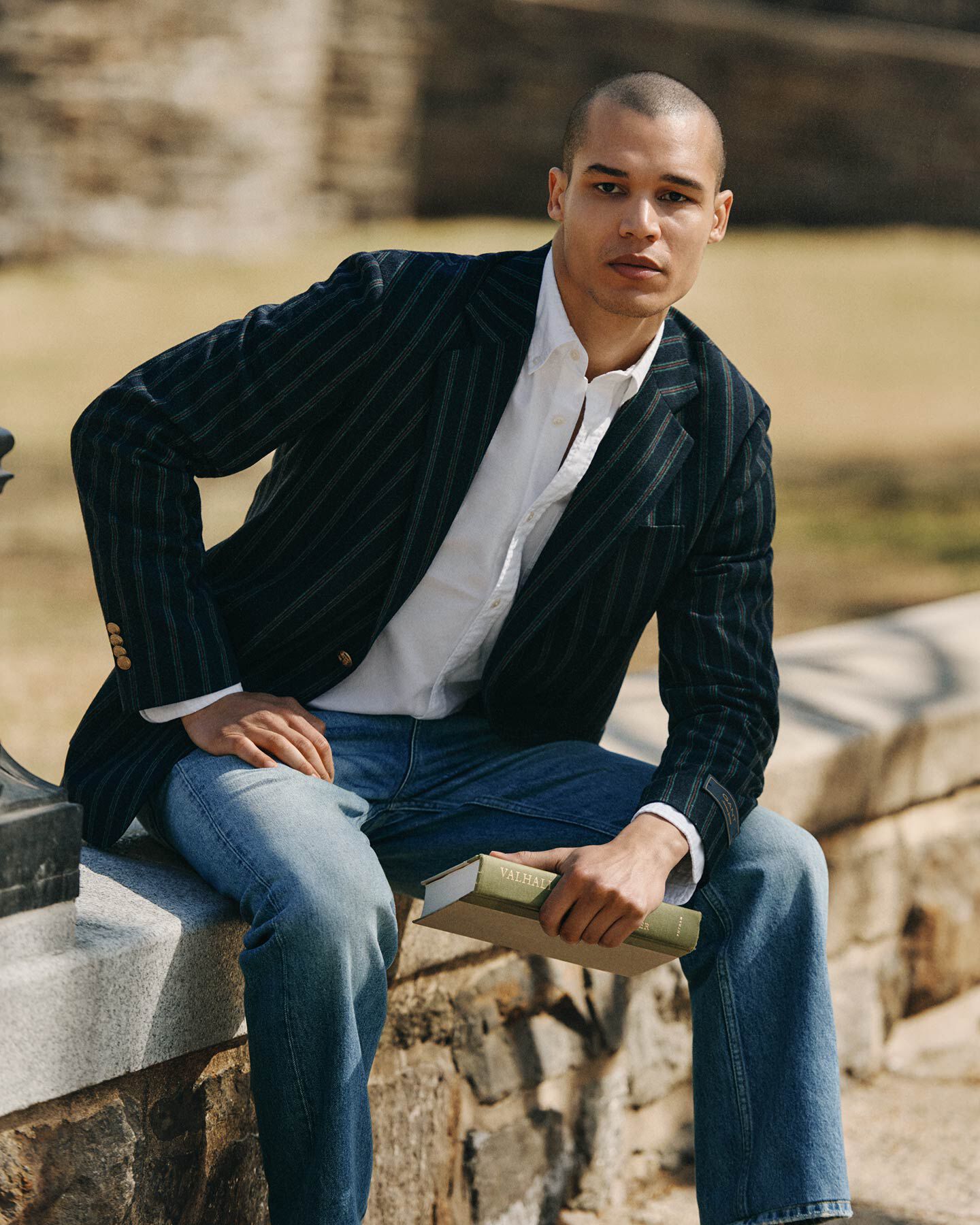 Man wearing Gant blue striped blazer and jeans sitting on a stone ledge.